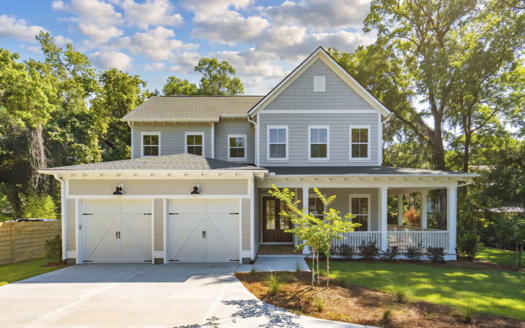 Two-story custom home with light gray siding, white trim, and a welcoming wraparound front porch. The exterior features a double garage with carriage-style doors, a gabled roofline with shingle accents, and mature trees surrounding the landscaped homesite.