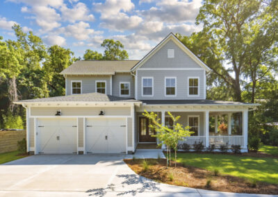Two-story custom home with light gray siding, white trim, and a welcoming wraparound front porch. The exterior features a double garage with carriage-style doors, a gabled roofline with shingle accents, and mature trees surrounding the landscaped homesite.