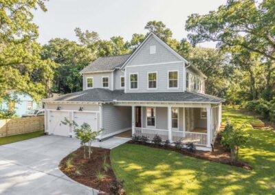 Two-story custom home with light gray siding, white trim, and a welcoming wraparound front porch. The exterior features a double garage with carriage-style doors, a gabled roofline with shingle accents, and mature trees surrounding the landscaped homesite.